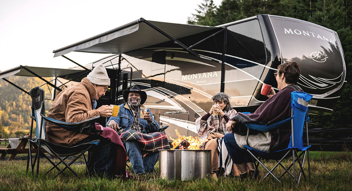 four friends around the campfire in front of a keystone montana legacy edition 5th wheel. Drinking coffee and laughing.