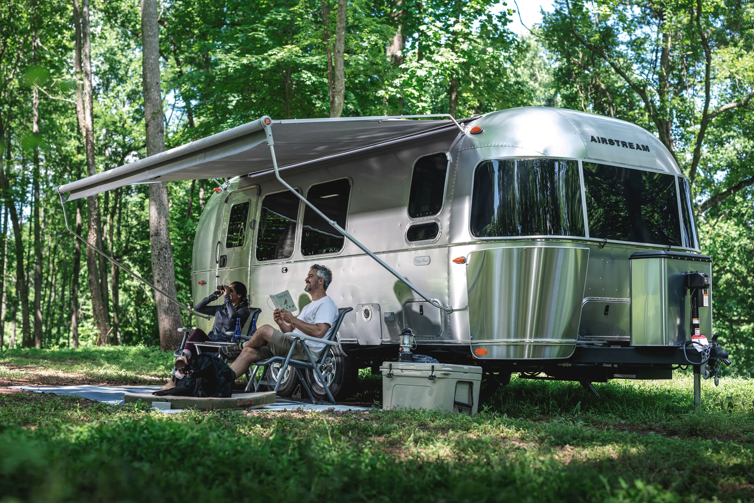 Couple relaxing under the awning of their Airstream Flying Cloud in the woods